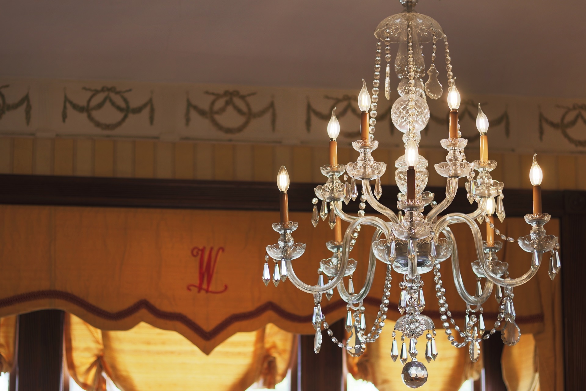 A crystal chandelier with lit candles hangs from the ceiling in a room with yellow curtains and patterned wallpaper.